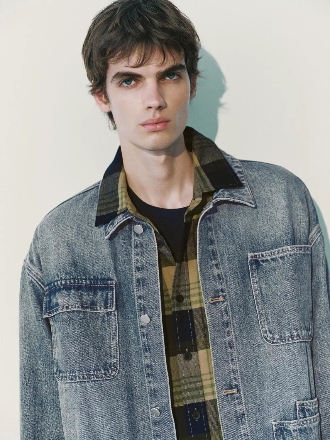 Denim jacket, light-wash blue with metal buttons and chest pockets over plaid shirt.