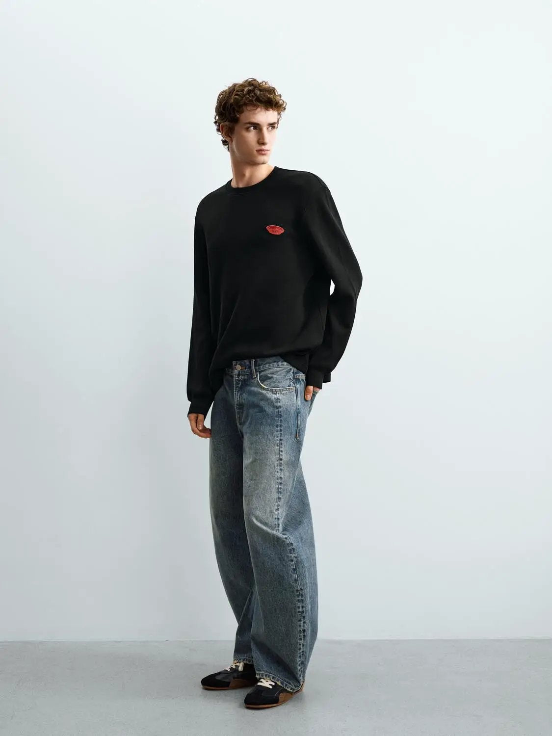 Sweatshirt, black long-sleeve with small red chest logo, relaxed fit on a model wearing blue denim jeans