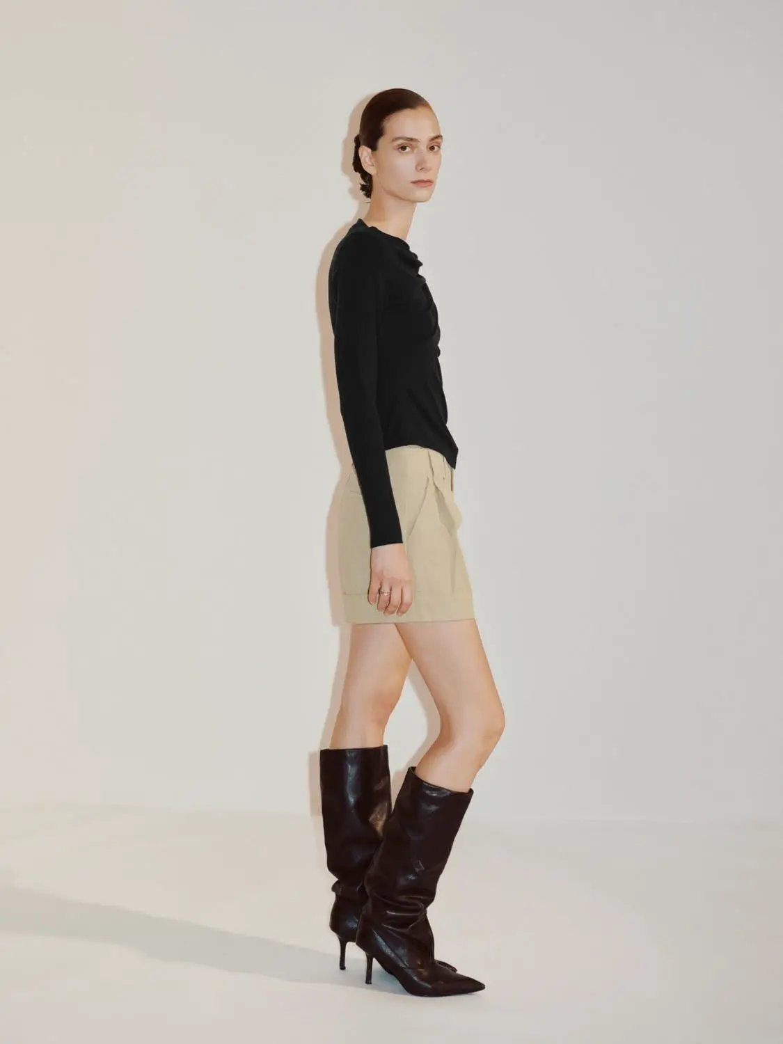Draped One-Shoulder Long Sleeve Blouse in black worn by a model; paired with beige shorts and tall black boots.
