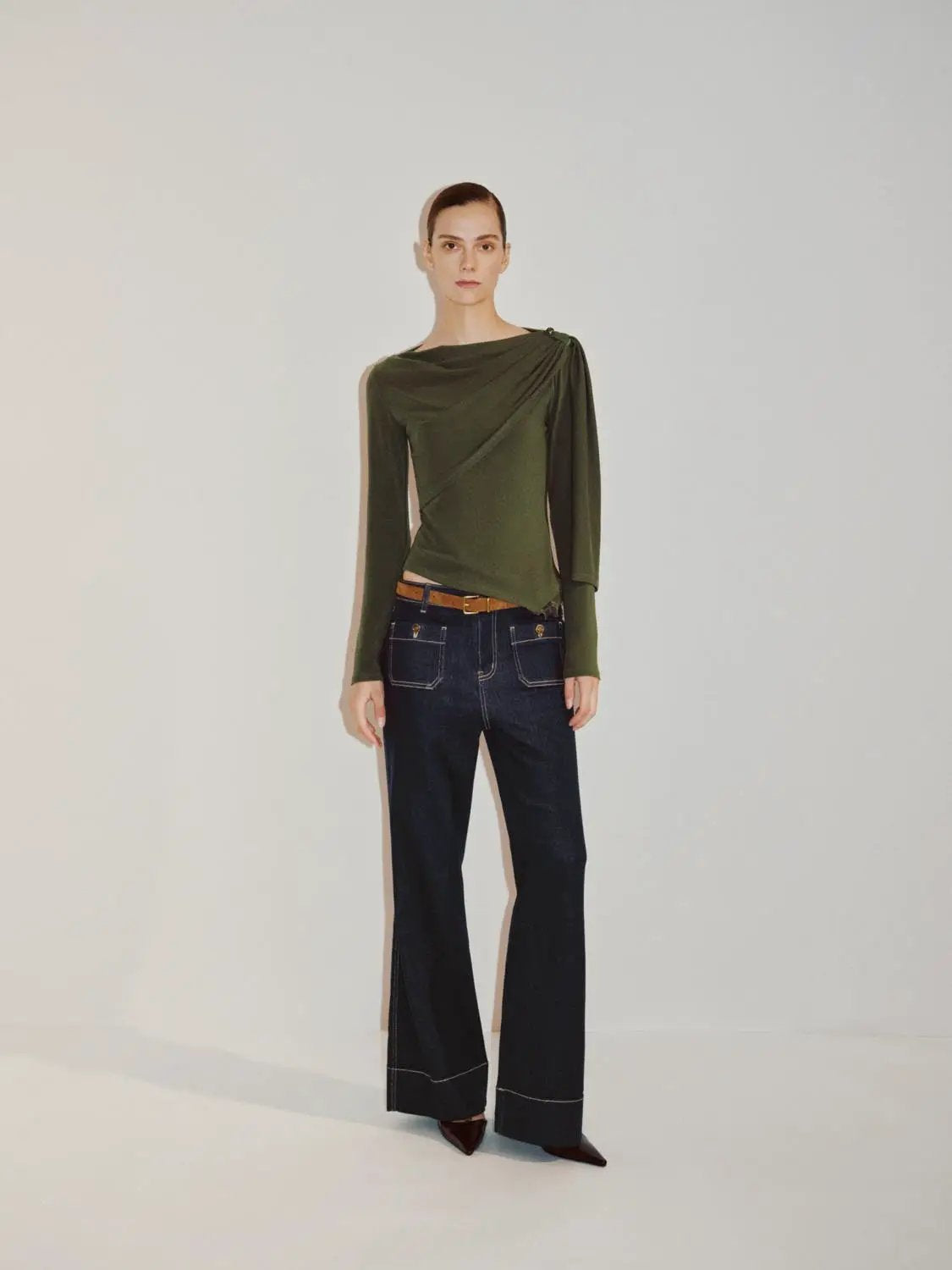 Draped one-shoulder long-sleeve blouse in olive green, with asymmetrical bodice; paired with dark wide-leg denim jeans.