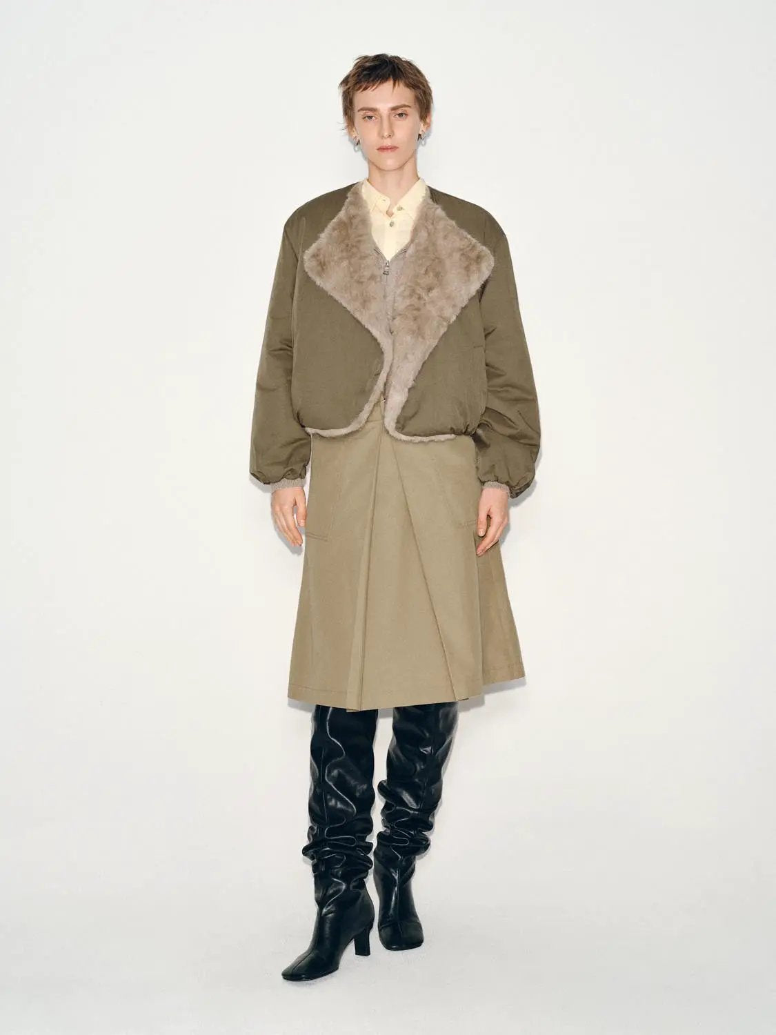 Pleated mid-rise belted A-line beige midi skirt on a model with an olive jacket and black knee-high boots.