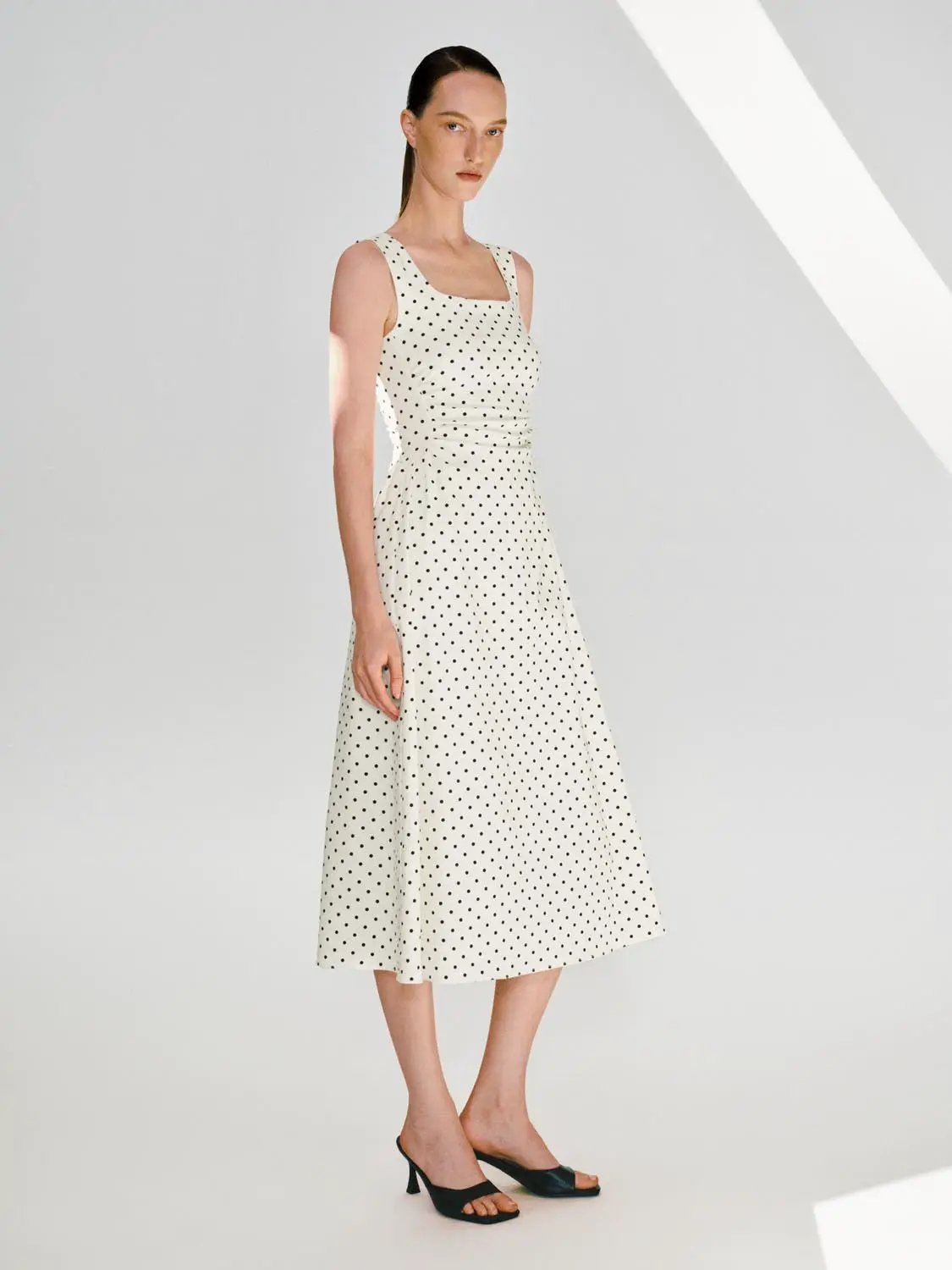 Polka dot knit A-line sleeveless dress, cream with black dots, midi length, model wearing black heels.