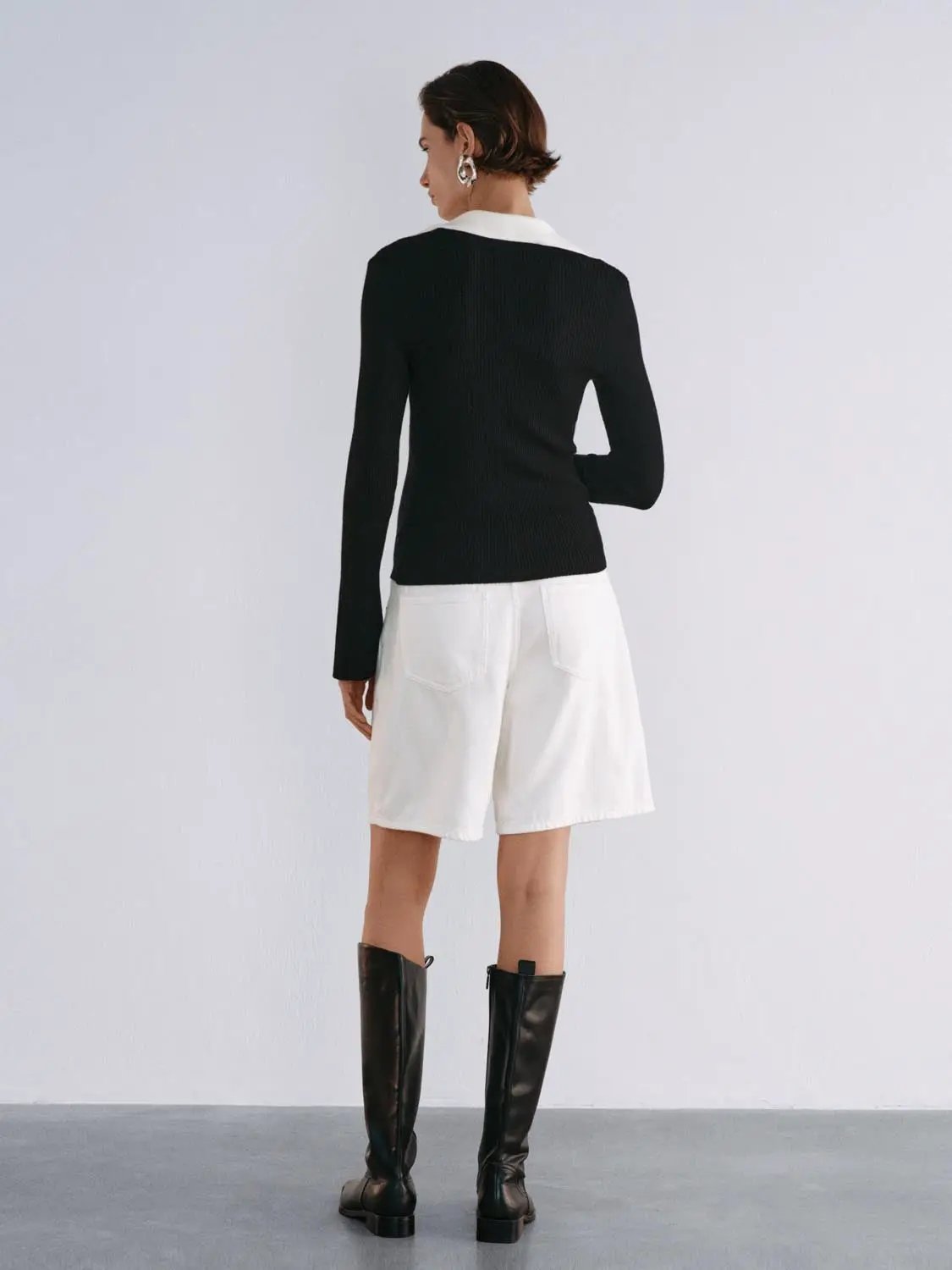 Knitted cardigan in black worn over white shirt, paired with white shorts and knee-high brown boots.