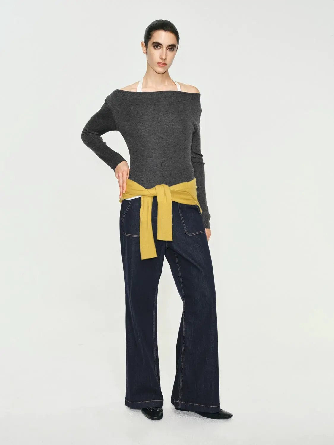 Fitted off-shoulder boat-neck knit top, dark grey; long-sleeve; mustard sweater tied at waist over dark denim jeans.