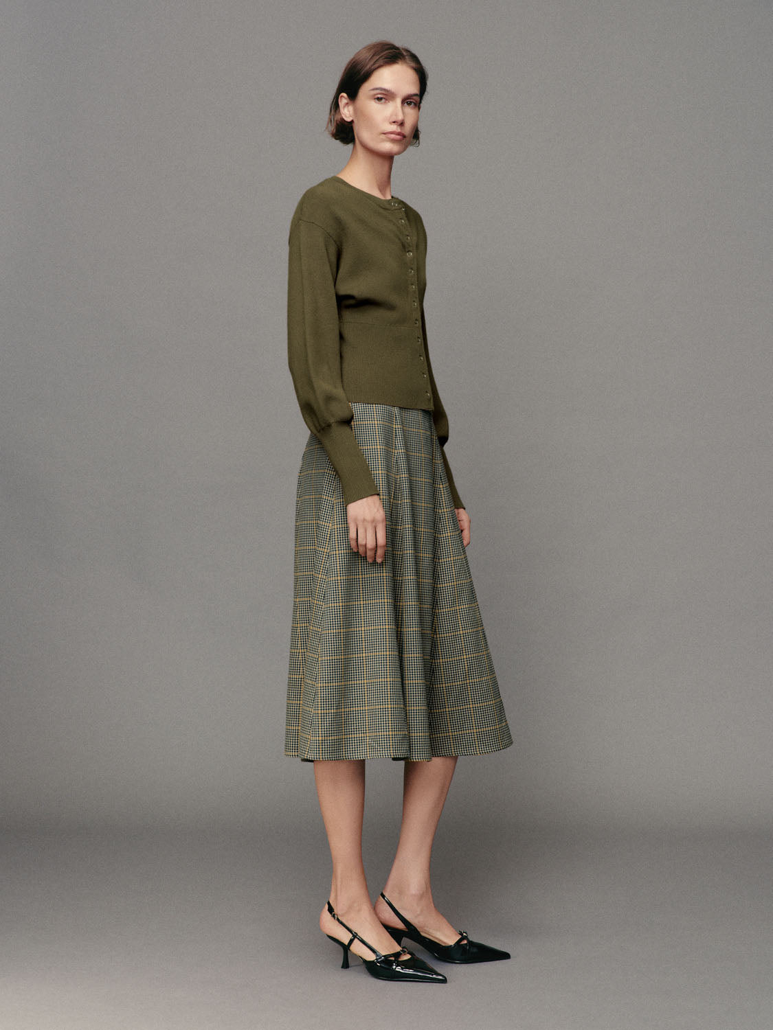 Olive green ribbed button-front cardigan with crew neck and long sleeves, paired with a plaid midi skirt.