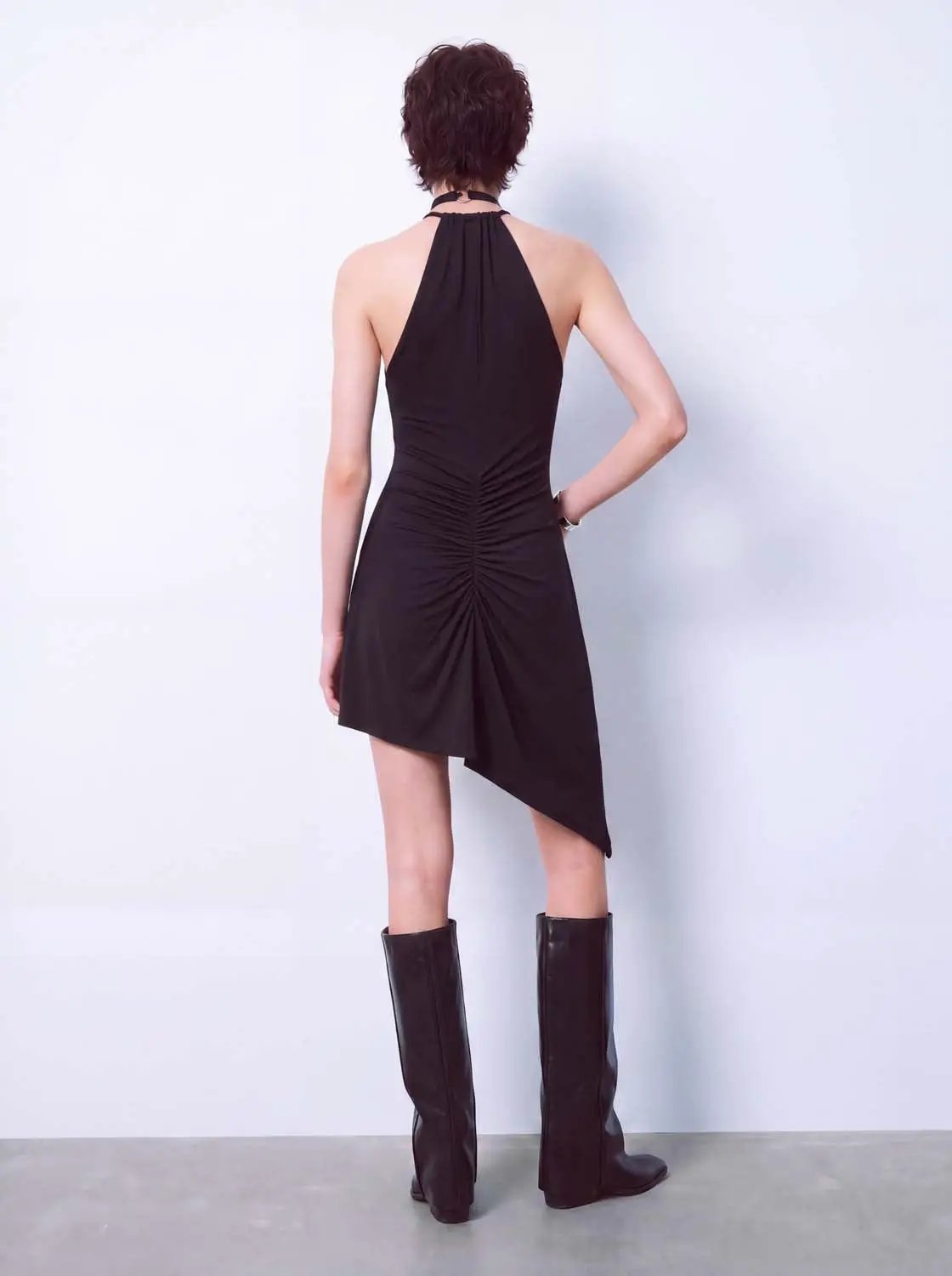 A-Line dress in black with halter neck, ruched back, and asymmetric hem, worn with knee-high boots.