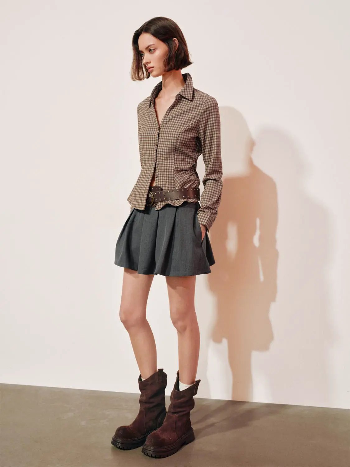 Button-front shirt in brown gingham worn by model, styled with a gray pleated mini skirt and brown boots.
