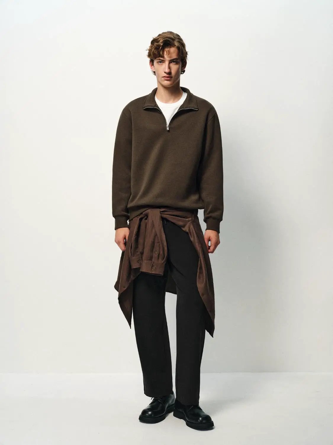 Sweatshirt in brown fleece with a half-zip collar; styled with dark pants and a brown shirt tied at the waist.