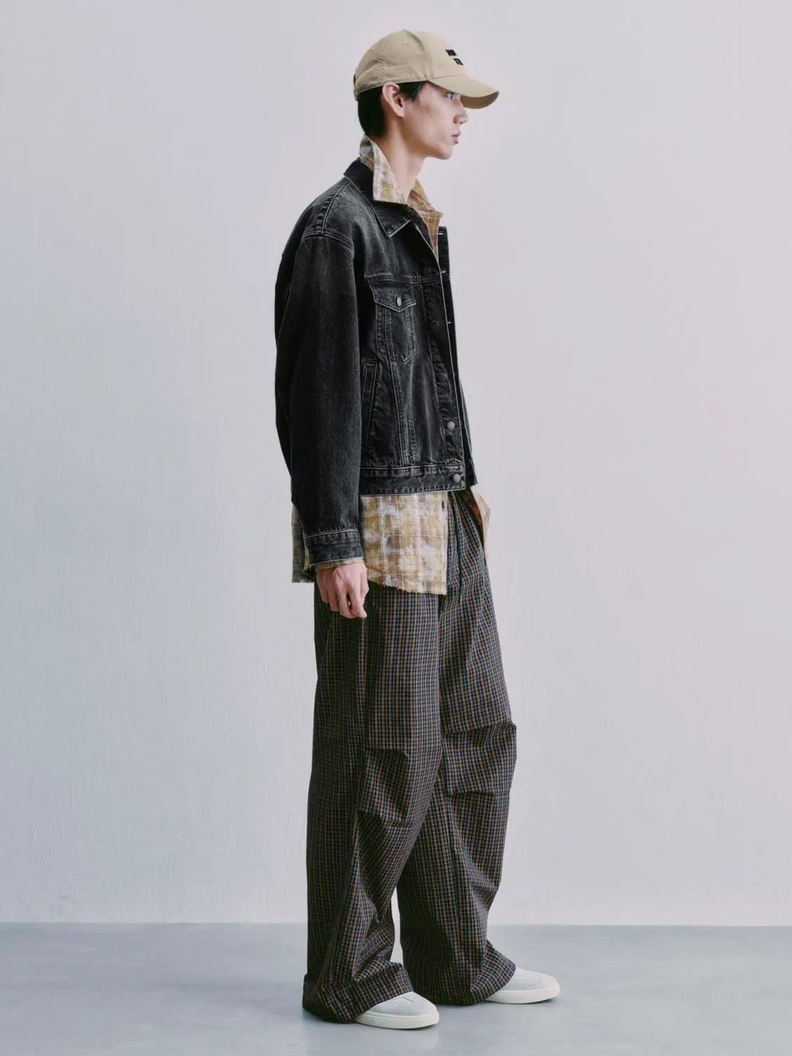Denim jacket in black wash worn over a beige plaid shirt, paired with brown plaid pants and a beige cap.