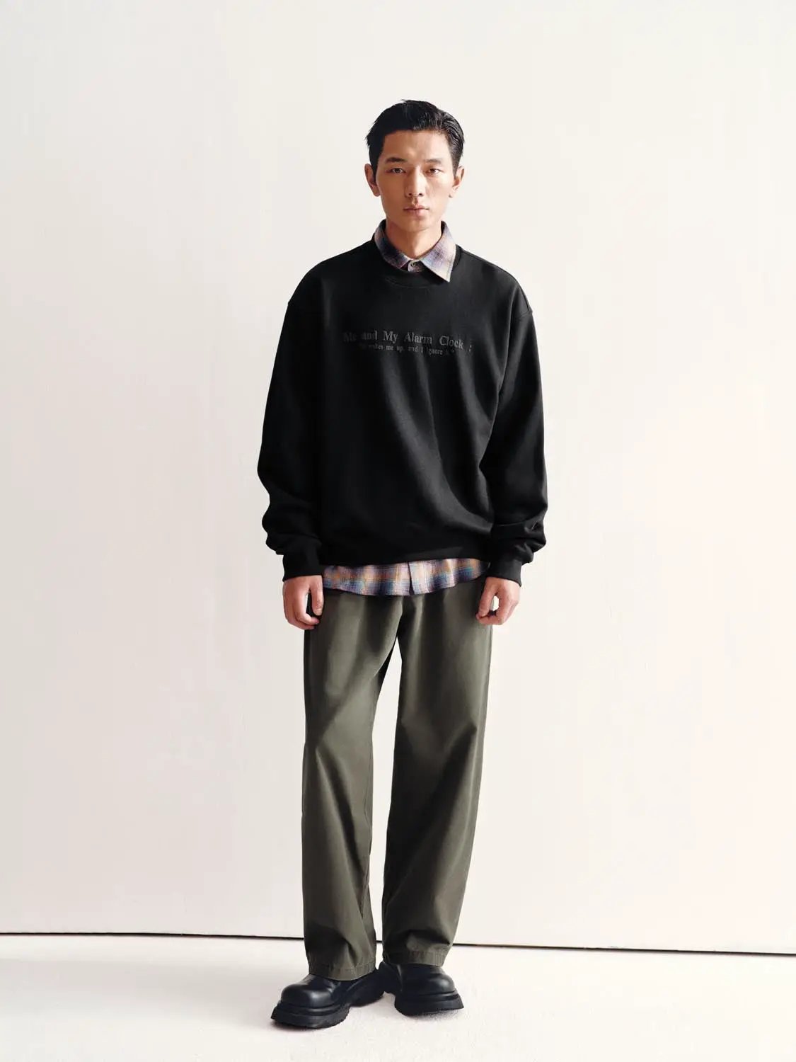 Sweatshirt black crewneck with subtle chest text; plaid shirt peeks out, paired with olive pants (3-piece outfit)