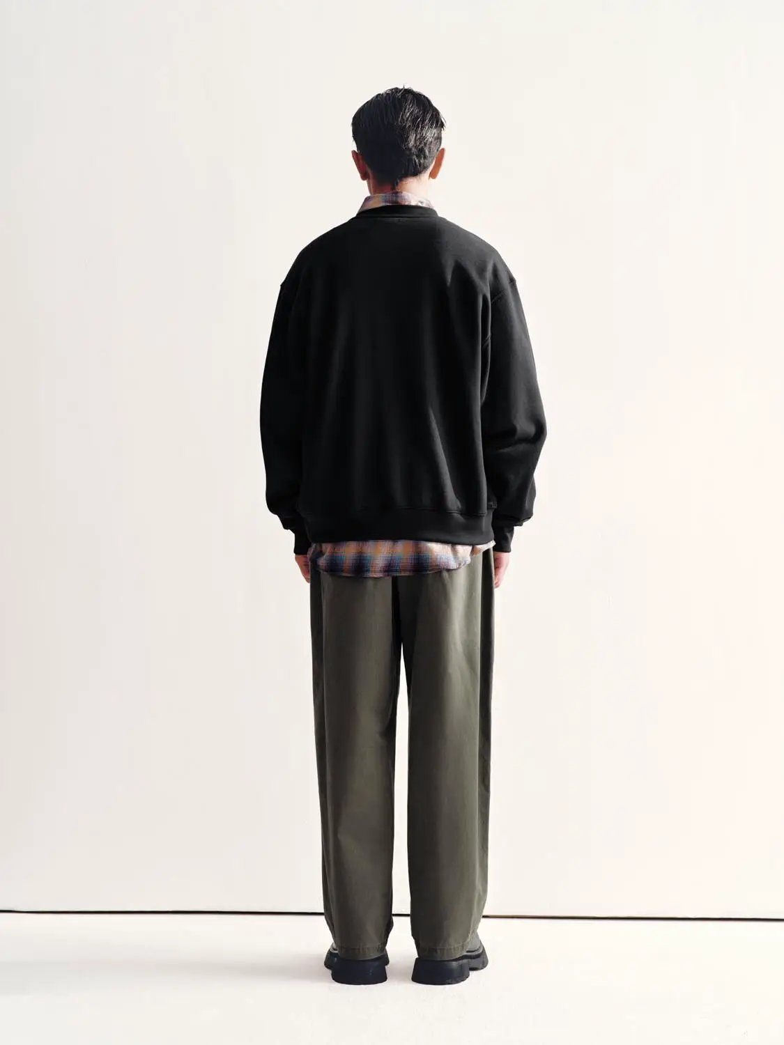 Sweatshirt, black, back view of model with plaid shirt peeking at the hem over olive pants.