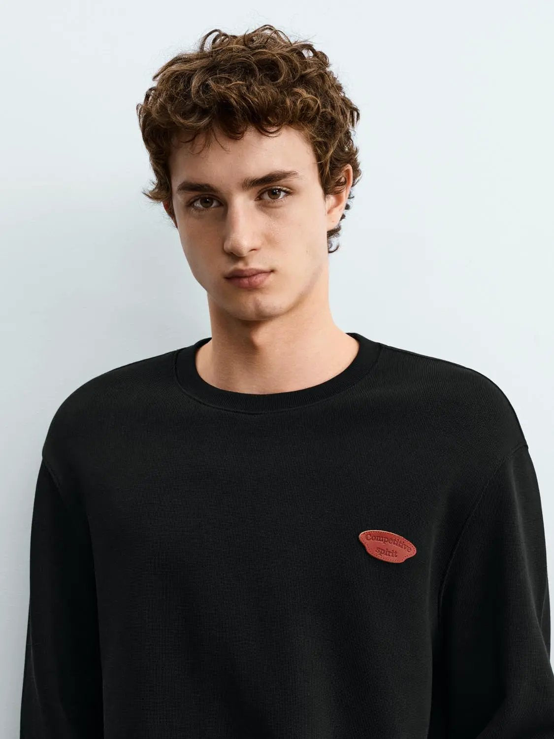 Sweatshirt black crewneck on male model with small red oval patch reading competitive spirit.