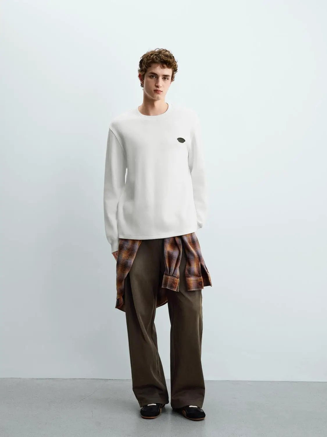 Sweatshirt white crewneck with a small chest logo worn by a model in brown pants with a plaid shirt tied around the waist.