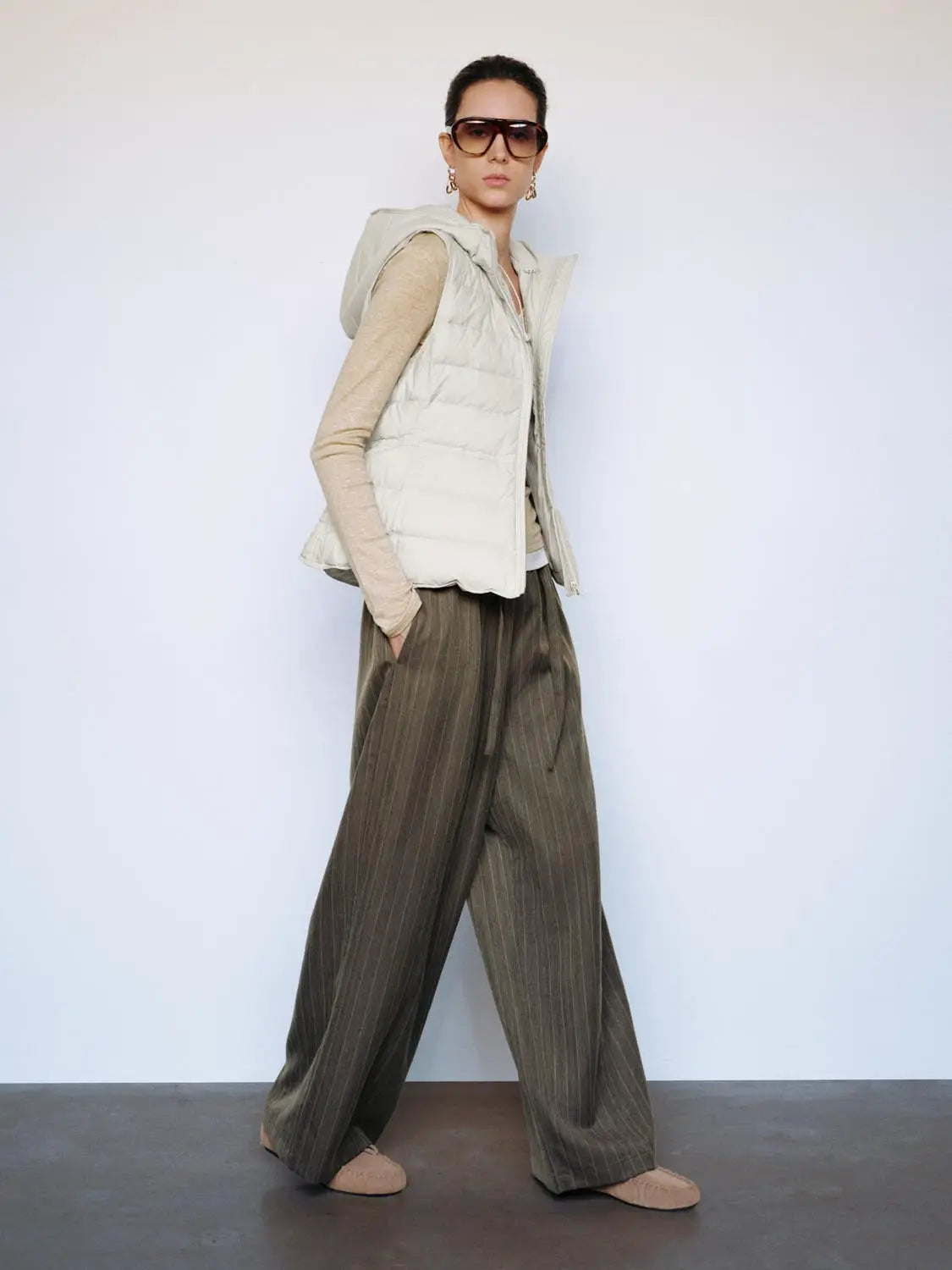 Down jacket vest in beige with hood from Urban Revivo Fashion, worn over a beige top and olive pinstripe wide-leg pants