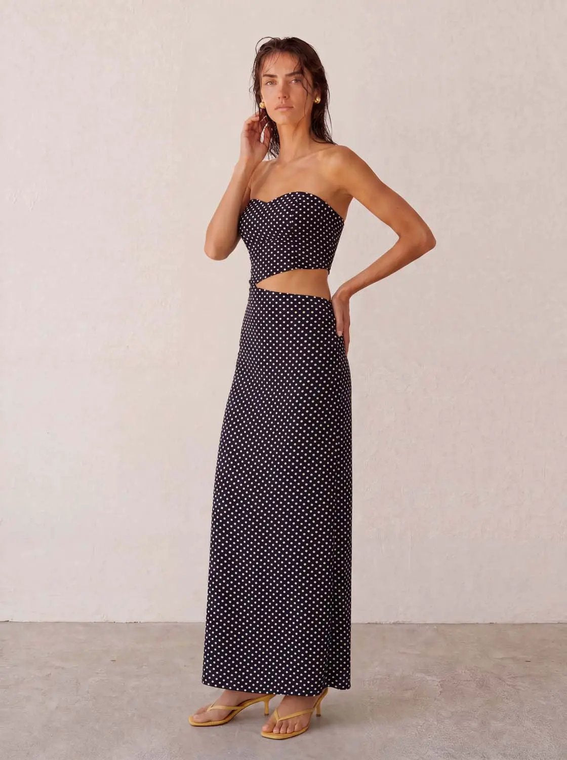 Dresses skinny-fit maxi in black with white polka dots, strapless with waist cutout, model wearing yellow sandals