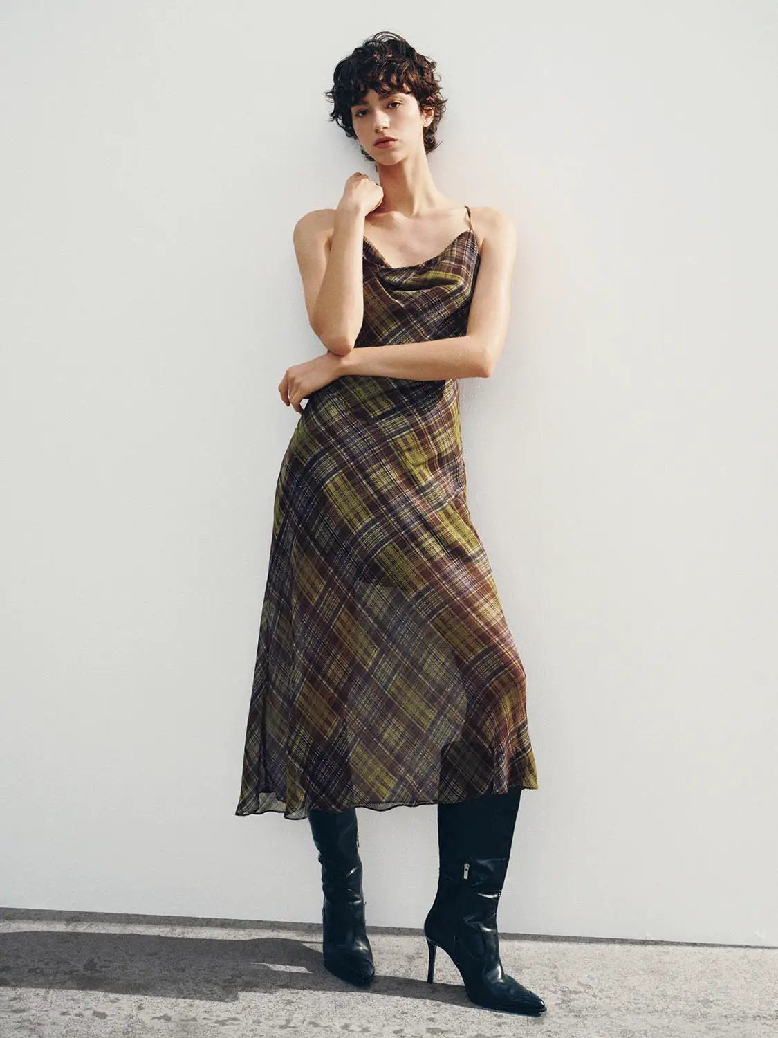 Plaid cowl-neck spaghetti-strap A-line dress on model; brown-olive plaid, knee-length, with black boots.