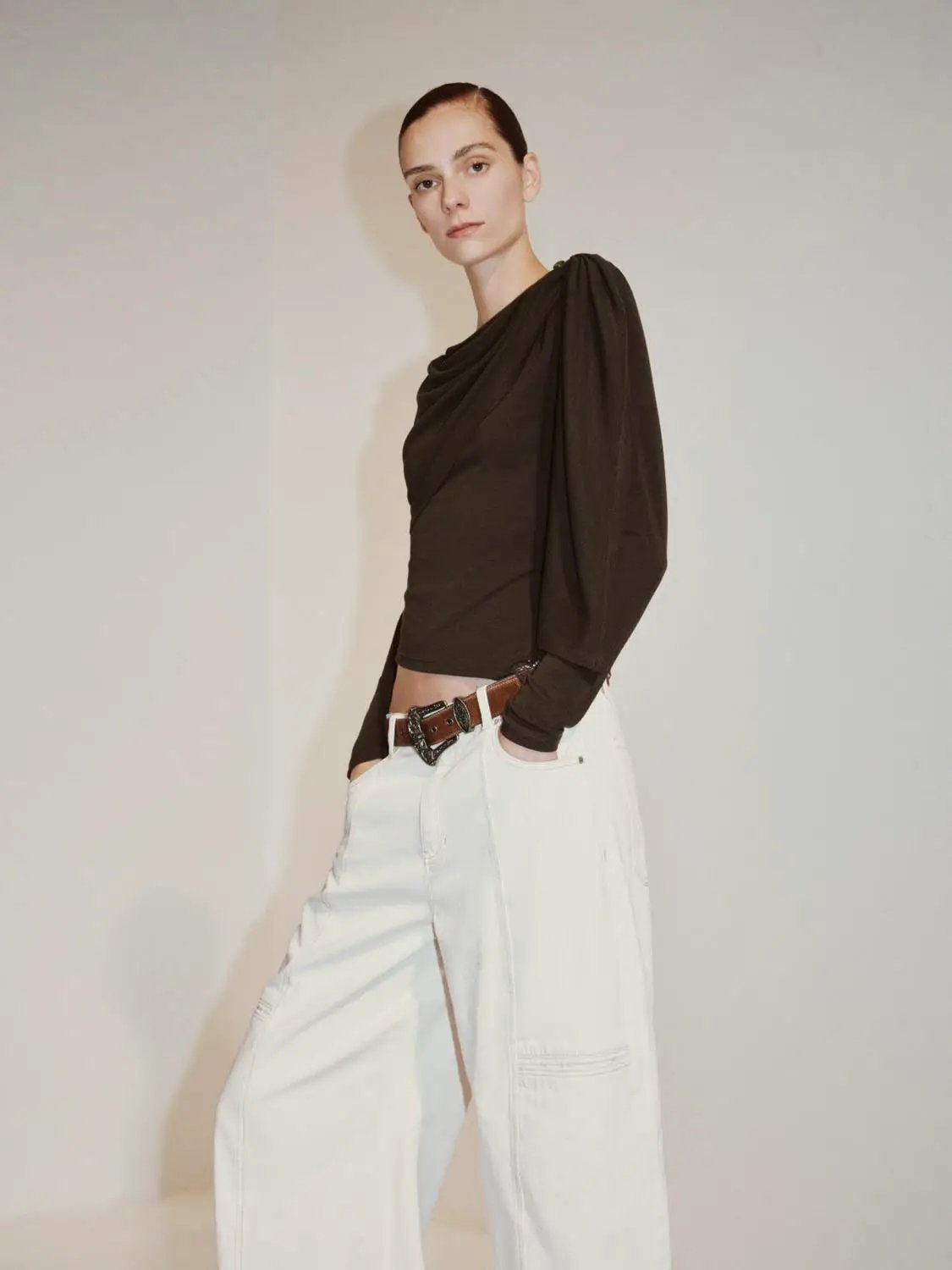 Draped one-shoulder long sleeve blouse in brown; paired with white pants.