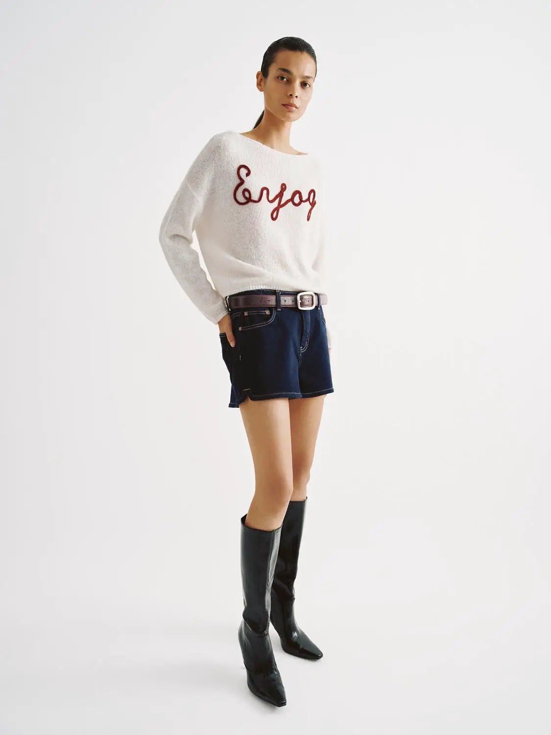 Knit lightweight white sweater with red script embroidery; model pairs it with dark denim shorts and tall boots