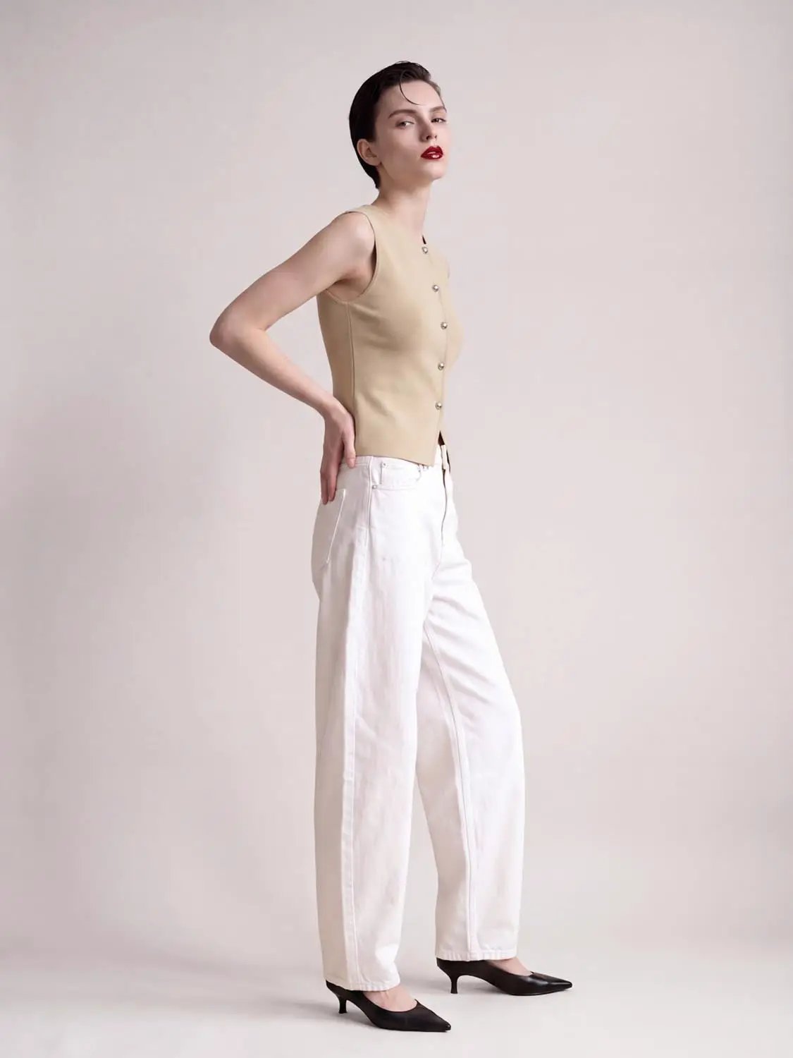 Round-neck button-front sleeveless cropped knit vest in beige with silver buttons, paired with white pants.