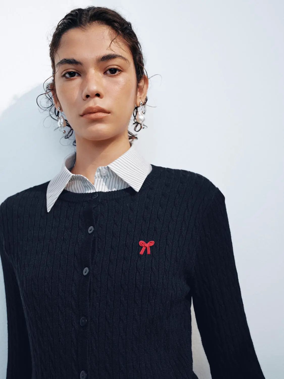 Black cable-knit cardigan with red bow emblem, button-front layered over a striped collared shirt.