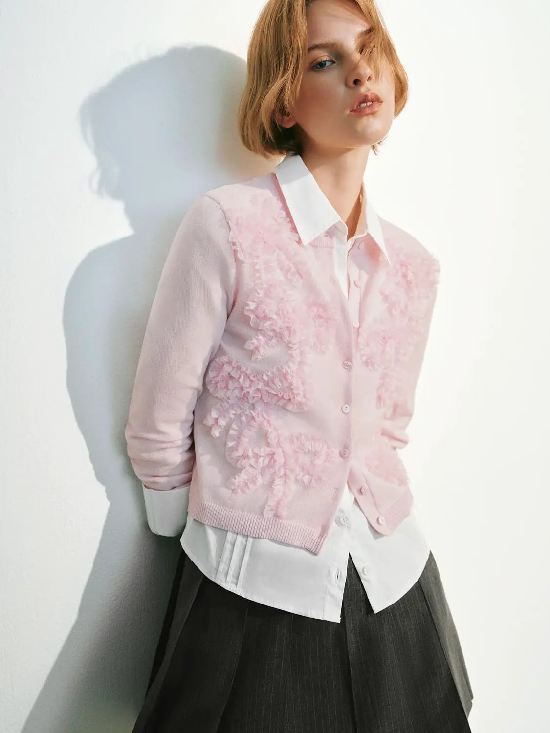 Knitted cardigan in pastel pink with ruffled floral texture, layered over a white shirt.