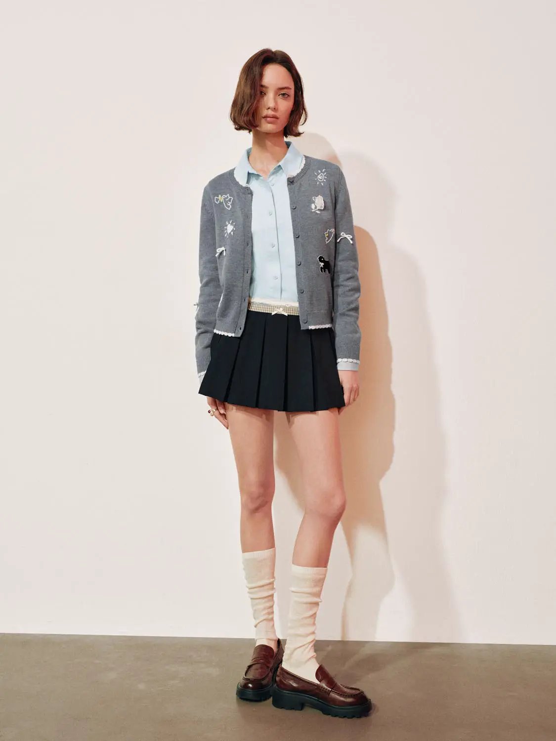 Graphic long sleeve ribbed knit cardigan in grey with embroidered patches worn over a light blue shirt and black pleated skirt