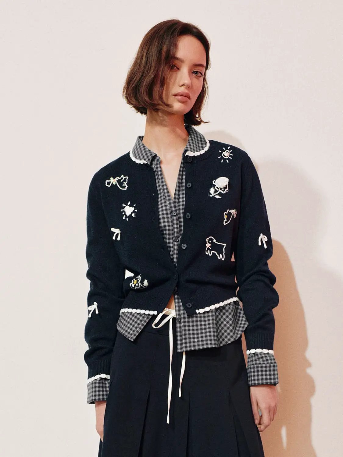 Graphic long sleeve ribbed knit cardigan in navy with white embroidered motifs, layered over a gray checkered shirt