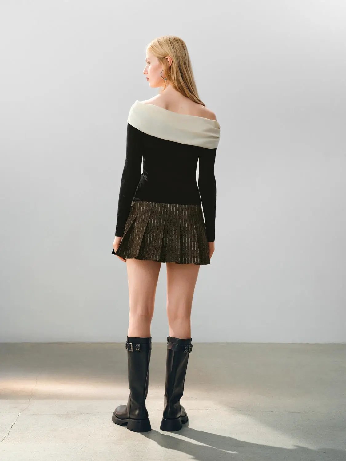 Knitted black off-shoulder top with a cream wide neckline, paired with a brown pinstriped pleated mini skirt.