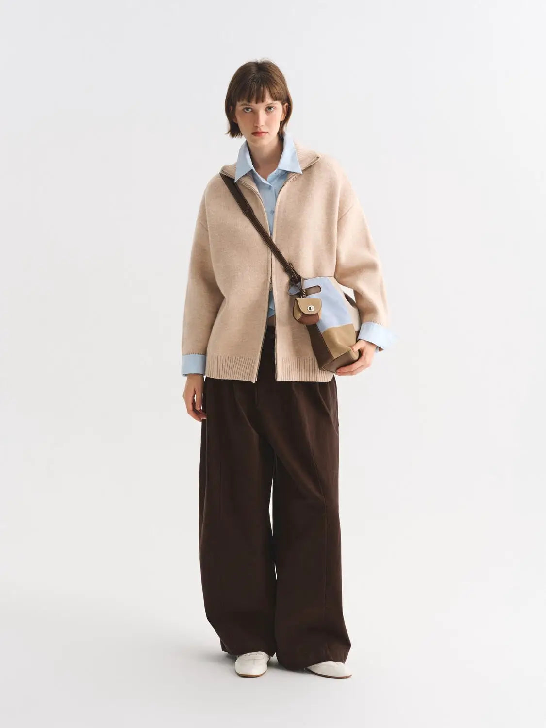 Knitted cardigan in beige, oversized fit, paired with a light blue shirt and brown wide-leg trousers.